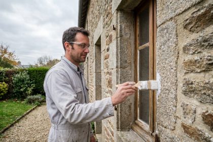Homme peignant le cadre de fenêtre d'une maison en pierre bretonne près de Rennes pendant le week-end
